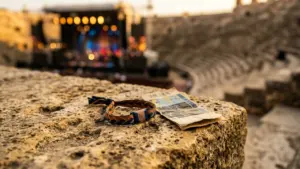Un bracelet de festival et un billet posés sur une pierre ancienne devant une scène.