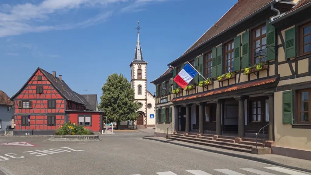 Une place de village traditionnelle française avec une mairie et un drapeau tricolore.