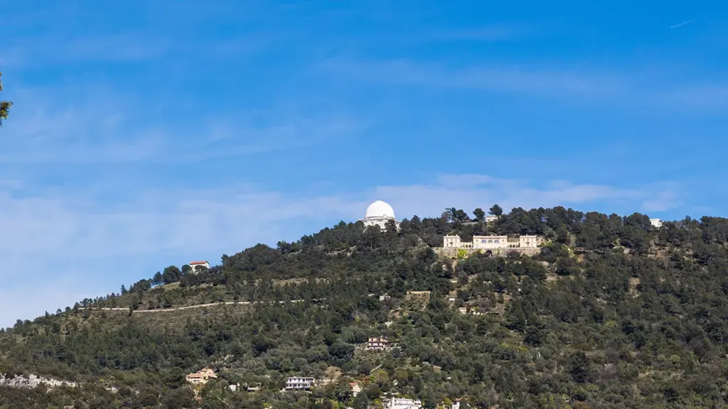 Un observatoire astronomique avec un dôme blanc situé au sommet d'une colline boisée ensoleillée.